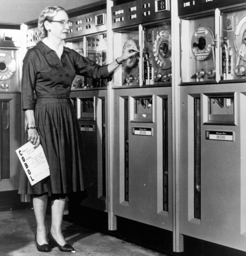 Photo of Grace Hopper with a COBOL manual standing in front of an old computer in 1952